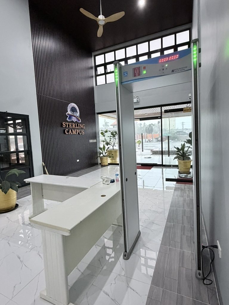 The main lobby of TAPS Sterling Campus with a white reception counter, walk-through security arch, and glass entrance doors beyond.
