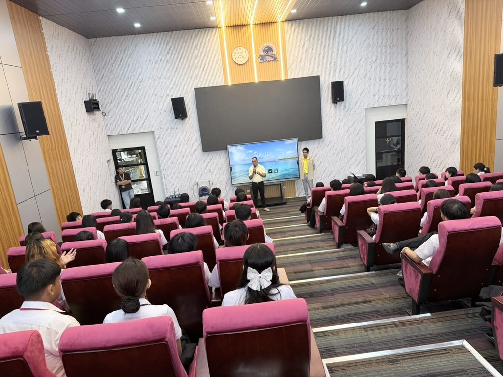 Arcinas Hall mid-program, the large stage screen lit with a presentation and the audience watching from tiered seats.