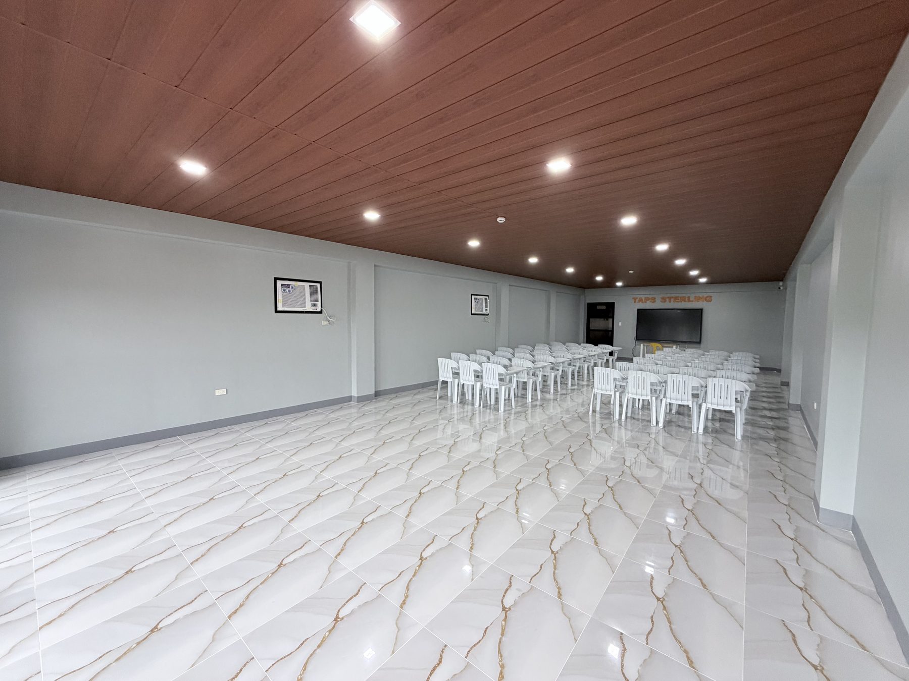 The seminar room seen from the doorway: rows of white stacking chairs on a marbled floor facing a wall-mounted display, under a timber-clad ceiling.
